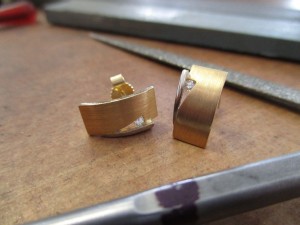 Goldene Ohrring mit einer kleinen, dreieckigen Kerbe geschlossen von einem silbernen Clip. In der Kerbe sitzt ein kleiner Schmuckstein.