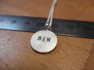 Ein kleiner, silberne Anhänger mit der Aufschrift "BEN" auf einem Lineal.
