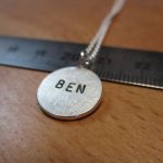 Ein kleiner, silberne Anhänger mit der Aufschrift "BEN" auf einem Lineal.