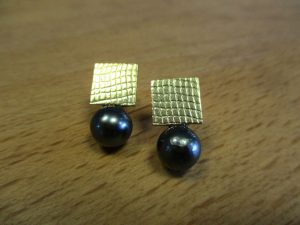Goldene, quadratische Ohrringe mit einer schuppenähnlichen Oberfläche. An der Unterseite sitzen schwarze Perlen.