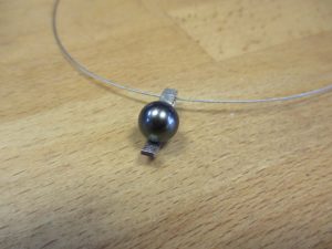 Silberner Anhänger in Form eines schmalen Streifen auf den eine dunkle, silberne Perle aufgesetzt ist.
